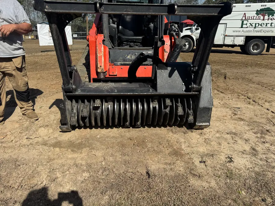Forestry mulching head on the front of a compact track loader, its drum lined with rows of bolt-on cutting teeth, an Austin Tree Experts chip truck parked behind it
