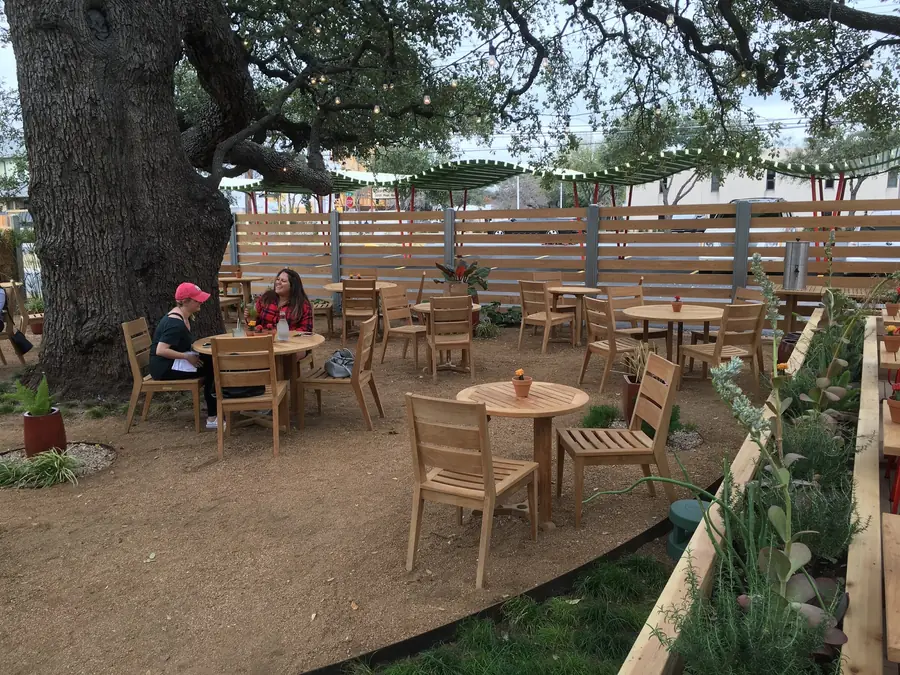 A gravel patio in use beneath a spreading canopy, tables running back towards a slatted timber fence