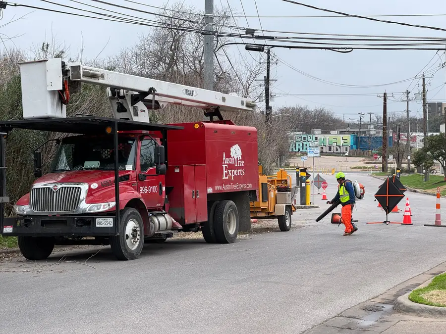Red Austin Tree Experts bucket truck at the curb with a chipper behind, crew in hi-vis on the road with traffic cones