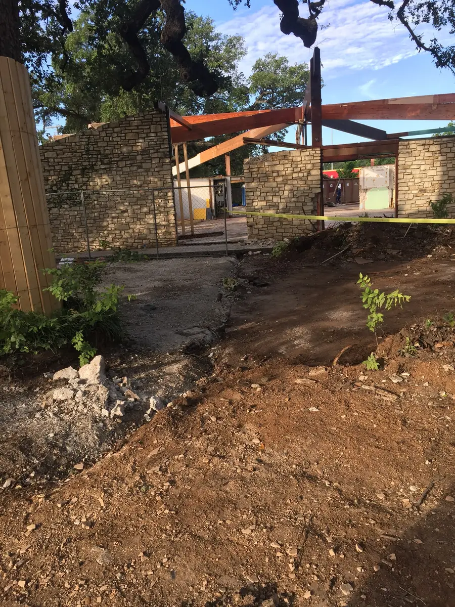 A construction site stripped back to bare soil with a steel frame rising beyond it, the ground around the retained tree left undisturbed